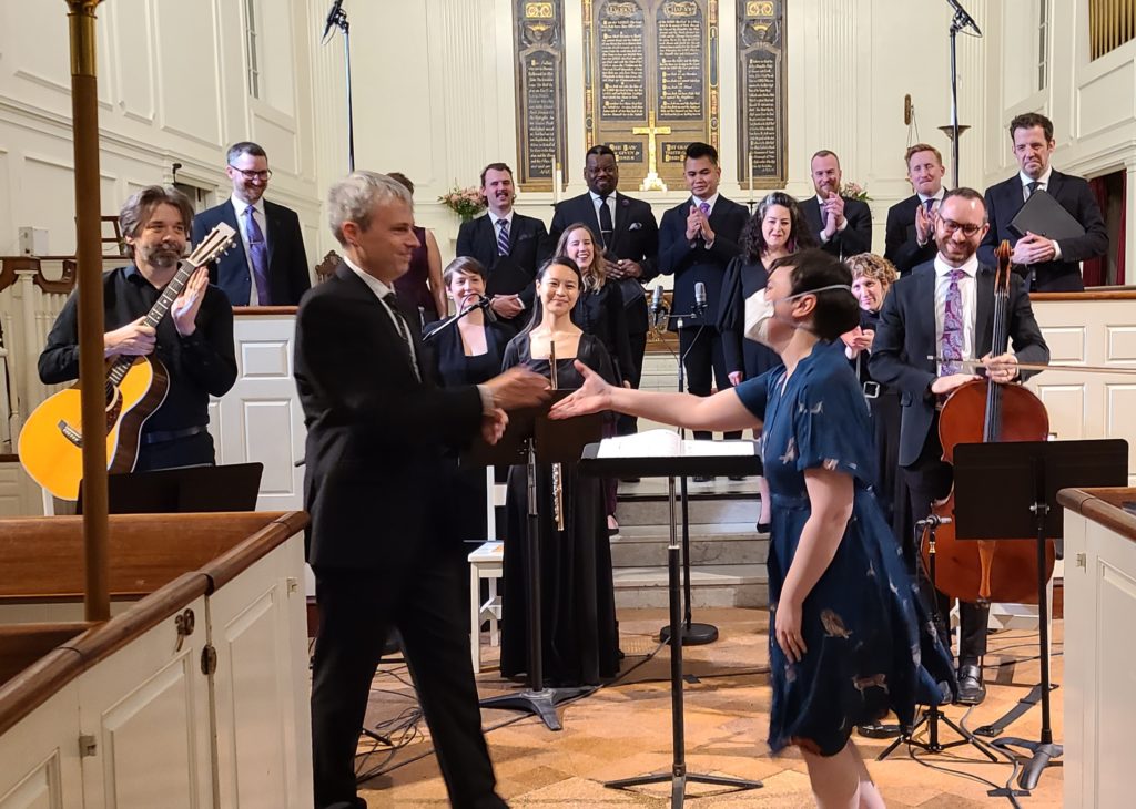 Nell reaches out to shake the hand of conductor Matthew Guard in front of the performing ensemble, standing for applause after their performance inside a historic church.