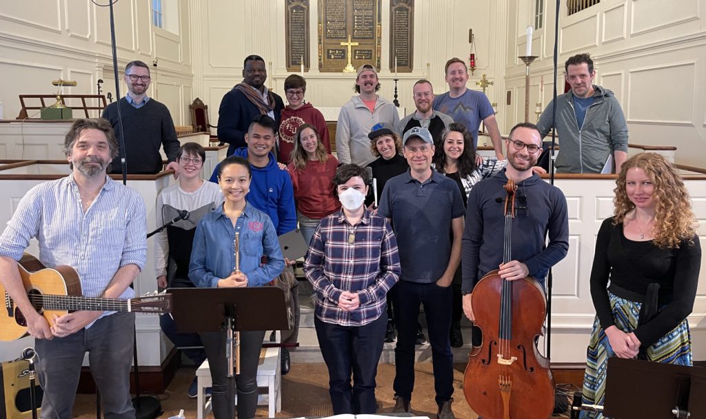 Nell poses with conductor Matthew Guard and an ensemble of 16 singers and instrumentalists, wearing casual clothes, during a recording session in a church venue.