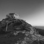 A black and white photo taken at the base of a staircase, looking up at a fire tower atop a mountain summit