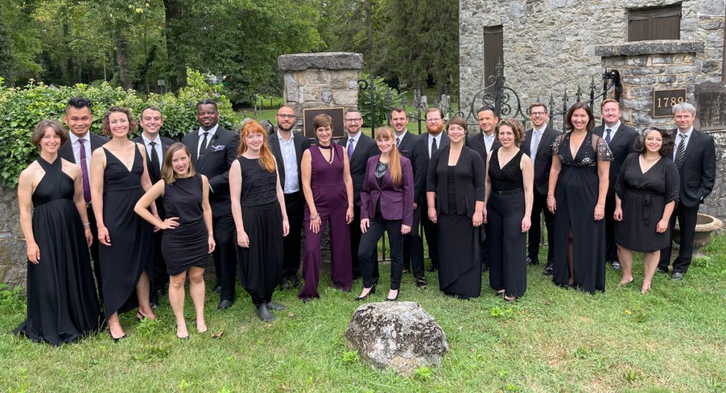 Photograph of Skylark Vocal Ensemble, wearing black and dark plum concert dress, standing in a semicircle and smiling toward the camera in a grassy outdoor setting