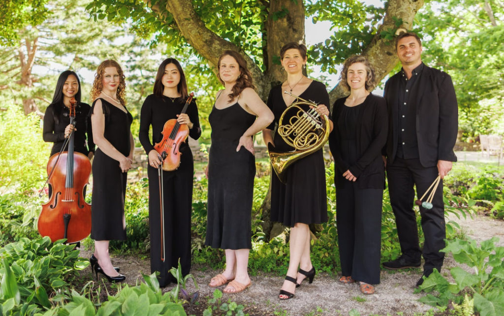 Juventas New Music Ensemble in concert dress, standing under a tree and holding their instruments as they face the camera