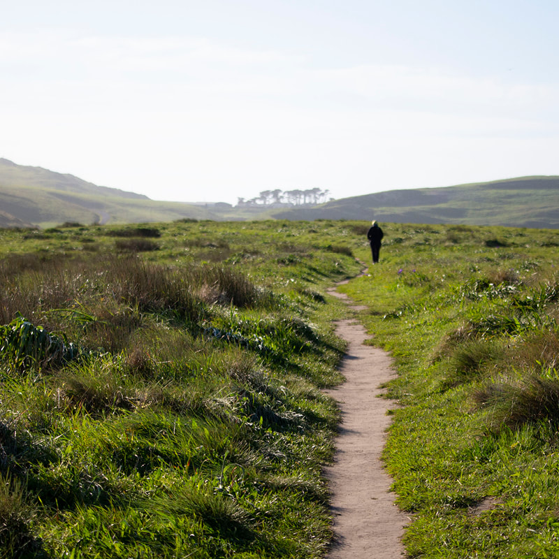 Photograph of dirt path through grassy hills, a figure walking in the distance