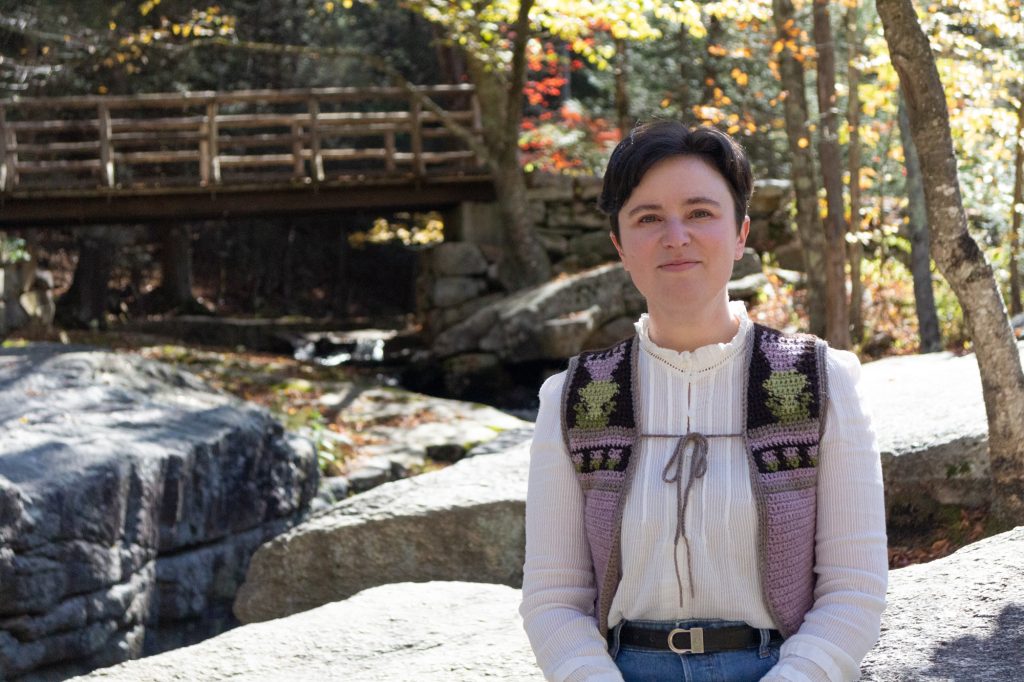 Nell Shaw Cohen sits on a rock in front of a creek in an autumnal forest.
